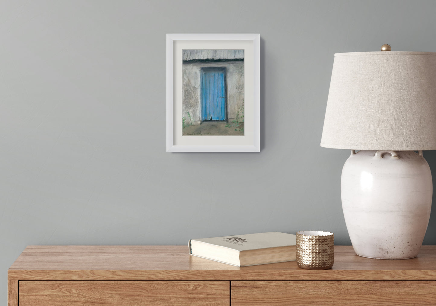 Framed artwork, lamp, and books on a wooden surface against a gray wall.